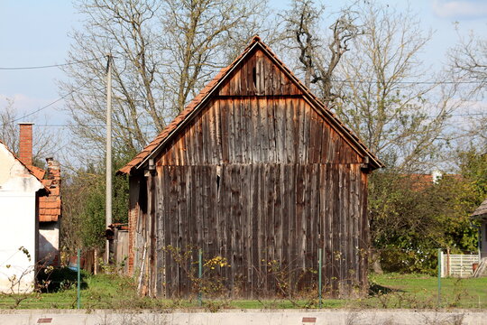 Side view of dilapidated old vintage rustic wooden barn with cracked wooden boards and leaning roof next to with abandoned family house ruins and tall trees without leaves in background on warm sunny 