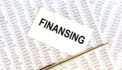 Text FINANSING on the page of a notepad lying on financial charts on the office desk. Near the calculator and marker. Business concept.