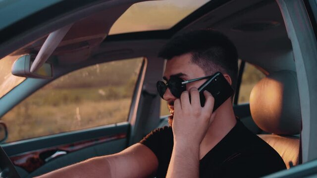 A Brunette Man With Sunglasses Is Talking On His Mobile Phone Behind The Wheel Of A Luxury Car. The Young Man Is Dressed In Light Summer Clothes. View From The Left Of The Driver