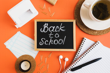 Naklejka premium Back to school flat lay: zeszyty, długopis, spinacze, dziurkacz, naklejki, filiżanka herbaty, świeczka i tablica na pomarańczowym tle