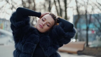 Portrait of a woman outdoors. Sexy girl in black fur coat looking on camera while holding her brown hair on the city park background. Slow motion.
