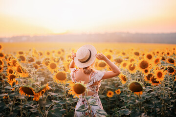 Beautiful summer nature amazing sunny sunset flowering field. Yellow orange sunflower flowers. Young modern woman turned away, enjoys harmony nature, rural relaxation. Girl touches straw hat on head