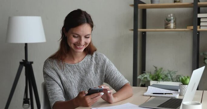 Millennial woman sitting at workplace desk distracted from study or work chatting with friend using smart phone receive pleasant message from boyfriend laughing. Modern distant communication, concept