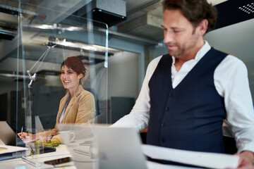 Smiling female entrepreneur seen through glass shield by colleague while working at creative office during COVID-19 pandemic