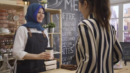 Cute Indonesian muslim waitress bringing hot coffee in paper cup and giving to client in restaurant. happy cafe employee in blue headscarf and waving hand greeting to customer in morning coffeehouse - Powered by Adobe