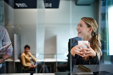 Cheerful beautiful young blond businesswoman holding cup while looking at colleague in office seen through glass shield