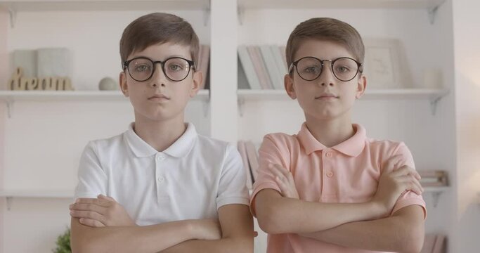 Twin brothers adjusting eyeglasses and crossing hands. Portrait of little Caucasian children posing at home. Smart kids looking at camera with serious facial expression.