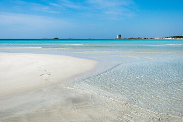 Spiaggia con sabbia bianca, mare trasparente e torre costiera sullo sfondo.
