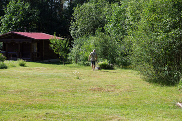 A white man mows the lawn of a dark-skinned hostess with an electric lawn mower against the background of trees and a wooden house...