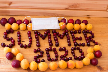 On the wooden surface of the cherry tree, the inscription SUMMER 2020 is laid out around it, plums with apricots, and a medical mask is placed on top.