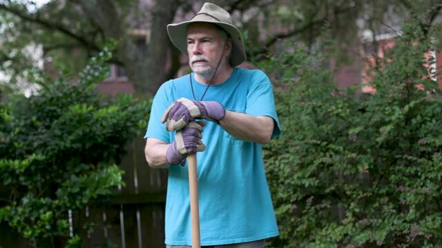 An older man doing yard work experiencing symptoms of heat exhaustion evident by profuse sweating and fatigue stops his digging for a moment.