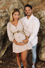 Pregnant couple touching their bellie in a forest and looking at the camera. Blonde pregnant woman with her brunette boyfriend. Blonde pregnant woman in a beige dress.