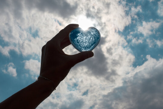 Hand holding up blue heart against a blue sky with white clouds. 