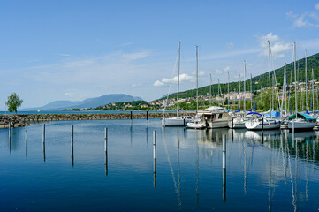 Small marina on the shores of Lake Neuchâtel. Very nice sunny weather. Rare clouds of fine weather in the blue sky. A few boats are moored. In the background, mountains and the houses of Neuchâtel