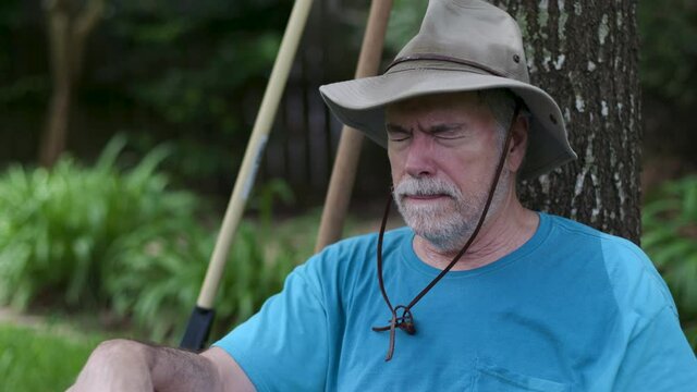 An older man sitting under a tree experiences symptoms of heat exhaustion or possible heatstroke evident by profuse sweating as well as extreme fatigue and confusion.
