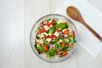 Fresh and healthy vegetables for salad in a glass bowl placed on white wooden surface.