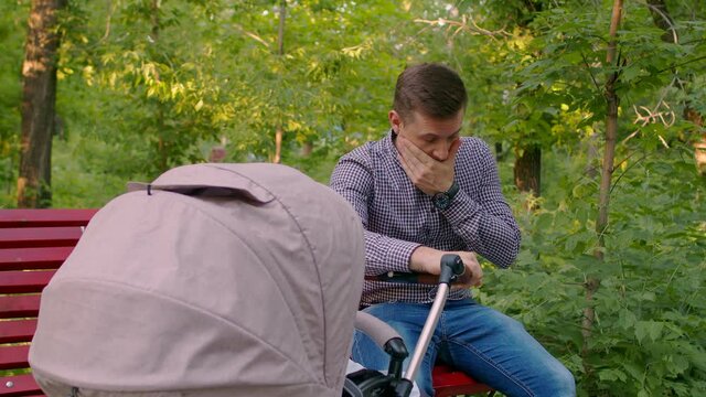 A tired father sits on a Park bench with his hand on a pram and rubs his eyes with his hand. A tired dad in a checked shirt and jeans walks with a baby in a stroller