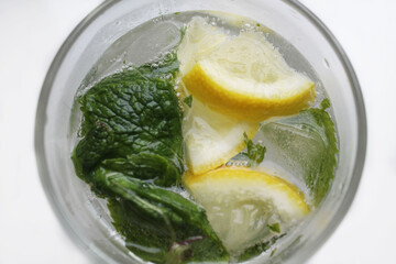 Round glass tumbler filled with water, close-up, top view. Fresh green mint petals and sliced ​​yellow lemon float on the surface of the water. Glass with a drink on a white background. Mojito.