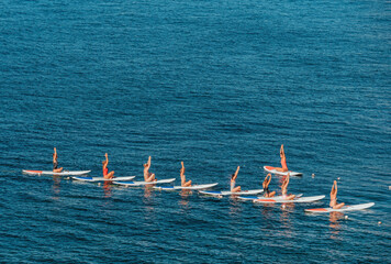 Group of young womens in swimsuits doing yoga on sup board in calm sea, early morning. Balanced...