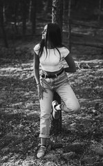 Young trendy pretty woman in moms jeans and a white top is leaning and holding her hand on a tree in the summer pine forest. Black and white photo