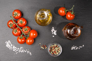 A  twig of cherry  tomatoes with some coarse salt grains, an oil and a vinegar jug and colorful pepper on a slate slab.