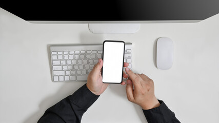 Top view of man’s hand holding smartphone on white table in home office with computer, keyboard, mouse and blank screen smartphone. concept work from home