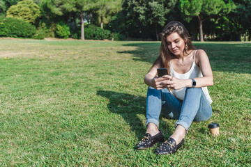 beautiful young caucasian woman sitting on the grass of an urban park texting with an smart phone