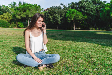 Naklejka na ściany i meble beautiful young caucasian woman sitting on the grass of an urban park talking with mobile phone
