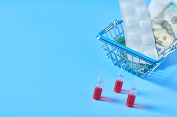 Red vaccine near pills and banknote of 100 dollars in market basket near glasses on blue background. Purchasing or sell of medicaments. Healthcare concept