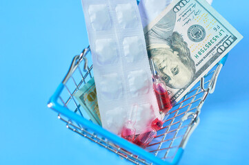 Red vaccine, pills and banknote of 100 dollars in market basket near glasses on blue background. Purchasing or sell of medicaments. Healthcare concept
