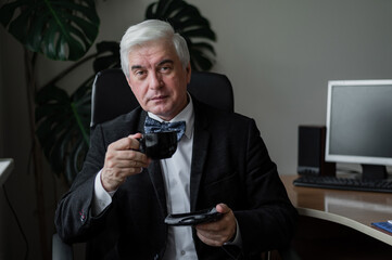 An attractive mature man in a suit and a bow tie is drinking coffee while sitting at his desk. Confident gray-haired male boss on a break in the office.