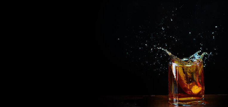 Ice cube dropped into a glass of whiskey with flying splashes on a black background. Stretched panoramic image