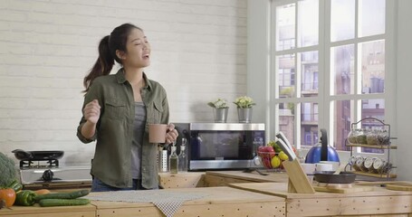 slow movement of cheerful asian chinese woman dancing and singing in kitchen at home in the morning. relax joyful lady drink coffee in apartment. happy young girl holding mug and moving body indoors - Powered by Adobe