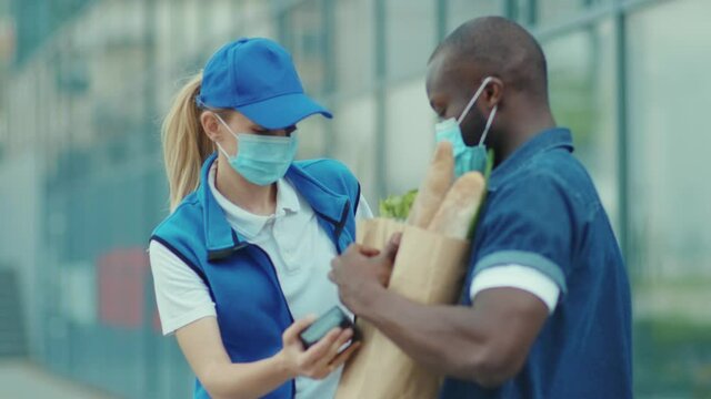 Portrait shot of young woman courier in blue hat and in medical mask give grocery packet to african american man client paying by contactless terminal payment worker outdoors slow motion