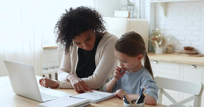 Adult afro american foster parent or babysitter helping little pretty caucasian girl preparing homework indoors. Caring biracial tutor giving private educational lesson to small attentive schoolgirl.