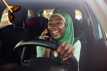 Obraz premium Muslim businesswoman on a business trip, driving a car and talking with smartphone. miling black muslim woman driving her vehicle. Happy muslim woman driving car