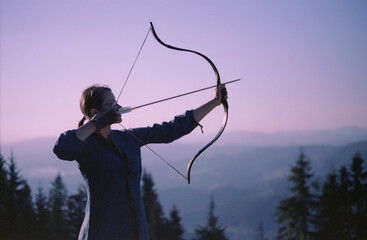 Woman shooting with a bow in the mountains. Young Caucasian female archer shooting with a bow at...