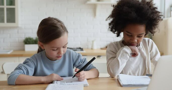 Happy cute little multiracial friends girls sitting at table, involved in entertaining math homework. Smiling small mixed race foster sisters discussing logic exercises, homeschooling study concept.