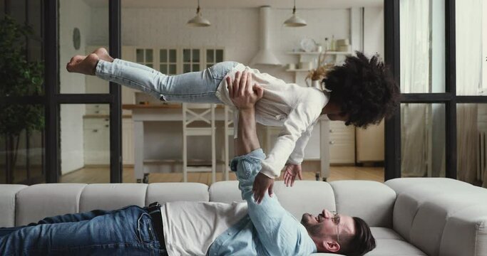 Smiling young caucasian father lying on sofa, holding on straight hands happy excited small african ethnicity adopted kid daughter, playing plane together indoors, interracial family hobby pastime.