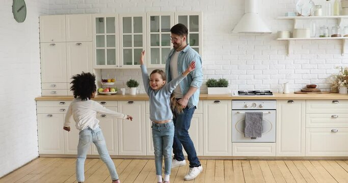 Full length overjoyed multiracial family dancing in modern kitchen, enjoying weekend time together. Happy young caucasian father having fun with energetic adopted mixed race children at home.