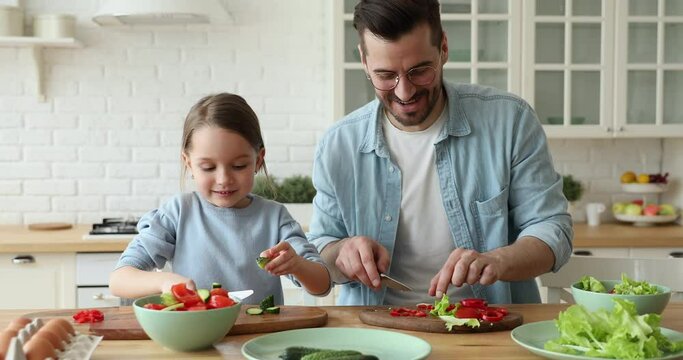 Adorable little preschool kid girl involved in cooking with affectionate caring father in modern kitchen. Happy young father preparing healthy vegetarian salad for lunch meal with cute daughter.
