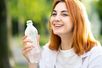 Young thirsty redhead woman drinking water from a bottle in summer park.
