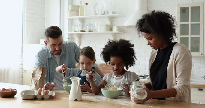 Little cute focused multiracial kids girl wearing aprons, learning making dough for pastry with affectionate loving mixed race parents, interracial family involved in hobby together in modern kitchen.