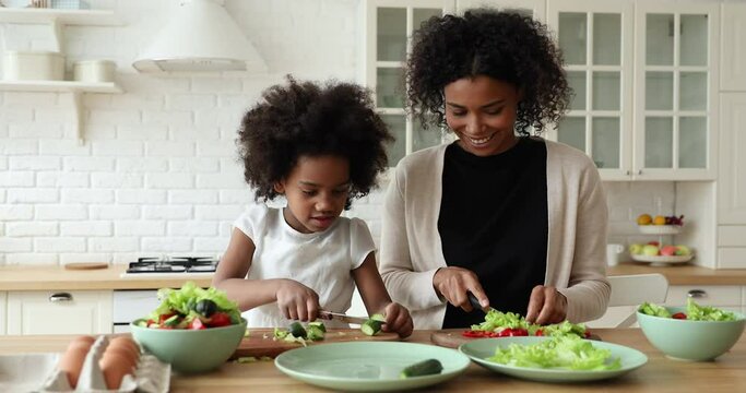 Little cute afro ethnicity kid girl helping biracial mommy preparing homemade healthy lunch in modern kitchen. Smiling two generations african american family enjoying cooking food together indoors.