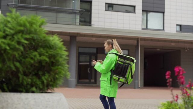 Tracking slow motion shot of teenage female food courier in green uniform and with thermal bag walking down street using navigation app on smartphone and calling doorbell to enter building