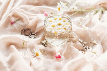 Fototapeta na wymiar Tender floral aesthetic. Daisy flowers in water in glass cup on background of soft beige fabric with wildflowers and jewelry. Creative summer image with space for text. Bohemian mood