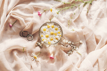 Obraz na płótnie Canvas Tender floral aesthetic. Daisy flowers in water in glass cup on background of soft beige fabric with wildflowers and jewelry. Creative summer image, flat lay. Bohemian mood