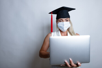 A beautiful graduate in a protective mask on her face and a graduation cap holds a laptop in her hand