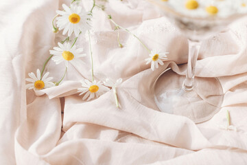 Daisy flowers and white wildflowers at wineglass stem on background of soft beige fabric. Tender floral aesthetic. Creative summer image. Bohemian mood