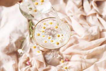 Pouring water with daisy flowers in modern wineglass on background of soft beige fabric with wildflowers. Tender floral aesthetic. Creative fresh summer drink. Bohemian mood